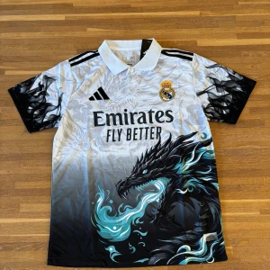 Real Madrid x Adidas draktröja M  | Unik design - Säljer en unik Real Madrid fotbollströja från Adidas i storlek M. Tröjan är vit med svart och turkos drakmotiv, har svarta Adidas-ränder på axlarna och klubbmärke broderat på bröstet. Tillverkad i lätt och ventilerande material – perfekt för match, träning eller streetwear.  ✔️ Lätt och andas ✔️ Kortärmad, regular fit ✔️ Aldrig använd  Storlek: M Skick: Ny / aldrig använd 