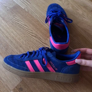 Adidas Spezial blå/rosa sneakers - Säljer ett par Adidas Spezial sneakers i blå mocka med rosa detaljer och klassiska tre ränder på sidan. Skorna har gummisula, snörning och retrovibbar. Perfekta för dig som vill sticka ut med färg och stil. Modellnamnet 'Spezial' syns på sidan.