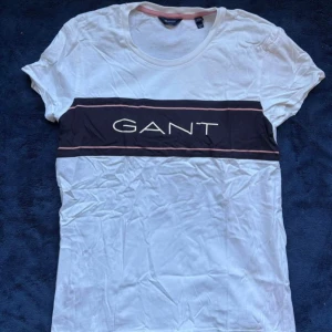 Vit GANT t-shirt med logga - Vit t-shirt från GANT. Klassisk rund halsringning och korta ärmar. T-shirten har en stilren design med tunna rosa linjer och är gjord i mjuk bomull för en skön känsla. Fick den i present men aldrig använd så den är som helt ny. Storleken är 170 cm (15 år) men sitter som en S. Fler bilder kan skickas om önskas och pris går att diskutera.