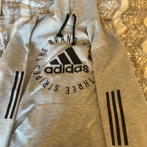 Grå Adidas hoodie med svart tryck - Snygg grå hoodie från Adidas med svart logga och text framtill. Tröjan har huva med svarta snören och de klassiska tre svarta ränderna på ärmarna. Perfekt för en sportig och avslappnad stil.