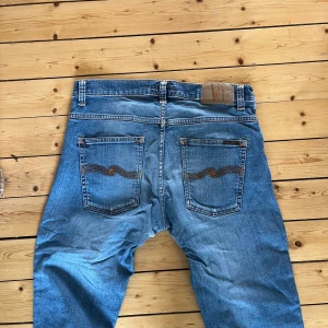 Blå jeans från Nudie Jeans - Säljer ett par klassiska blå jeans från Nudie Jeans med snygga slitningar och kontrastsömmar. Jeansen har raka ben, fem fickor och Nudie-loggan på bakfickan. Perfekta för dig som gillar en avslappnad och tidlös stil.