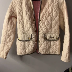 Beige  Barbour jacka -  beige jacka från Barbour med grå kantband och guldiga tryckknappar. Jackan har två stora fickor framtill med Barbour-logga jackan har inga tacken på användning