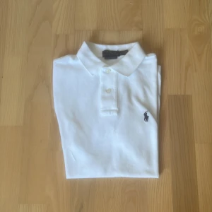 Vit pikétröja från Ralph Lauren - Klassisk vit pikétröja från Ralph Lauren med broderad logga på bröstet. Modellen är Custom Slim Fit med korta ärmar, krage och knäppning med två knappar. Tillverkad i mjuk bomull som känns skön mot huden. Perfekt för en clean och stilren look. Och är knappt använd