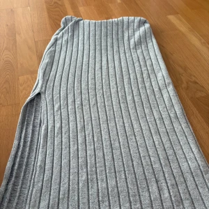 Grå ribbad långkjol i stickat material - Mysig grå långkjol i stickat material med ribbad struktur. Kjolen har en rak passform och är perfekt för dig som gillar en enkel men stilren look. Den är mjuk och följsam, vilket gör den superbekväm att bära hela dagen.