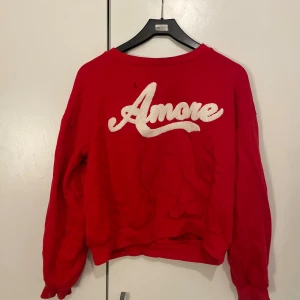 Röd sweatshirt med Amore-tryck - Cool röd sweatshirt med stort vitt Amore-tryck framtill. Tröjan har en avslappnad passform, rund halsringning och långa ärmar. Perfekt för dig som vill sticka ut med ett statement-plagg och gillar streetstyle. Storlek 158-164