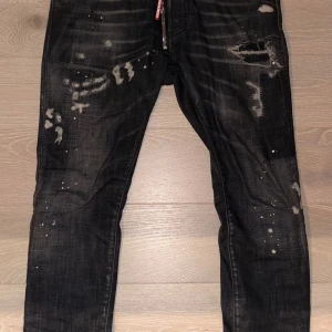 Svarta Dsquared2  Jeans med slitningar - Svarta Dsquared2  Jeans med coola slitningar, färgstänk och distressed detaljer. Jeansen har en röd dragkedja med Dsquared2-logga och klassisk femficksdesign. Snygg, köpt på Nk kvitto finns. Vid fler fårgor eller bilder skriv 