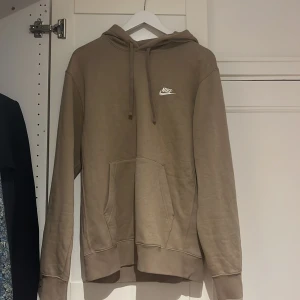 Beige Nike hoodie med broderad logga - Säljer en beige hoodie från Nike med broderad vit logga på bröstet. Tröjan har huva med snörning, en stor ficka och långa ärmar. Perfekt för chill dagar eller när du vill ha en clean och enkel stil.