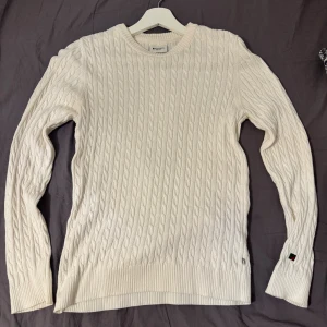 Vit kabelstickad tröja redgreen  - Vit kabelstickad tröja från redgreen med rund halsringning och diskret logga på ärmslutet. Tröjan har lång ärm och ribbade muddar för en klassisk look. Perfekt för lager-på-lager och snygg till både jeans och kjol.