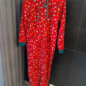 Julig röd fleeceonesie från Esmara - Supermysig röd fleeceonesie från Esmara med juligt HO HO HO-mönster, polkagrisar och färgglada detaljer. Grön huva och muddar, dragkedja framtill och långa ärmar. Perfekt för att maxa julkänslan hemma!