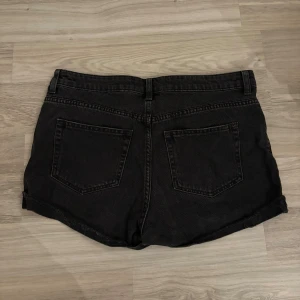 Svarta jeansshorts - Svarta jeansshorts med medelhög midja. Fyra knappar framtill, fickor bak och fram. Snygg vintagekänsla och perfekt till sommaren eller med strumpbyxor på hösten. Shortsen är i storlek 40 men passar som S också. 