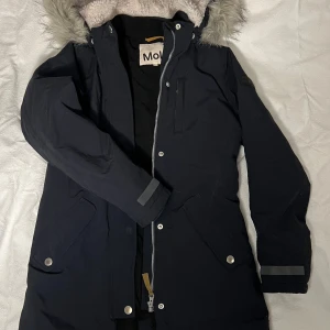 Mörk marinblå parkasjacka med fuskpäls från Molo - Marinblå parkasjacka från Molo i storlek 164. Jackan har huva med mjuk fuskpäls fästa med tryckknappar. Jackan har både dragkedja och tryckknappar framtill samt stora fickor med fleecetyg. Innerficka finns. Luvan är avtagbar, fodrad i mjuk ljusbiege fuskpäls. Materialet är återvunnen nylon och polyester med 3M Thinsulate-isolering. Perfekt för kalla dagar och snyggt urban look. Plagget är sparsamt använt och tvättat endast 1 ggn. Den är som ny! Nypris ca: 1400 kr. Säljes för 700 kr.