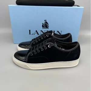 Lanvins - Snygga svarta sneakers från Lanvin med vit platt sula, ovandel i mocka och detaljer i skinn. Tån är i blankt lackskinn som ger en lyxig touch. Klassisk snörning och rund tå. Perfekta för dig som gillar stilrena och exklusiva sneakers.