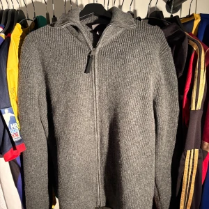 Grå stickad kofta med dragkedja - Mysig grå stickad kofta med hel dragkedja framtill. Tröjan har ribbad struktur och långa ärmar, perfekt för kyliga dagar. Enkel och stilren design som funkar till det mesta. 