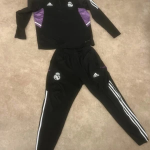 Real Madrid svart träningströja Adidas - Svart långärmad träningströja från Adidas med Real Madrid-logga och vita ränder på ärmarna. Snygga lila detaljer på sidorna och halv dragkedja vid halsen. Perfekt för fotbollsträning eller chill. Matchande byxor finns också.