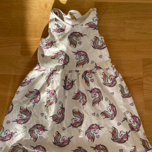 Vit enhörningsklänning H&M 110/116 - Supersöt vit klänning från H&M i storlek 110/116 med färgglatt enhörningsmönster och små hjärtan. Ärmlös modell med rund hals och utställd kjol. Perfekt för dig som älskar unicorns och pastellfärger!