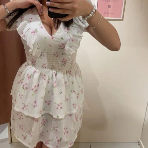 Vit blommig volangklänning Gina Tricot - Superfin kort klänning från Gina Tricot i vitt med rosa blommor och gröna blad. V-ringad med volangdetaljer vid axlar och byst, smockad midja och dubbla volanger i kjolen. Använd endast 1 gång ! Nypris: 399 kr