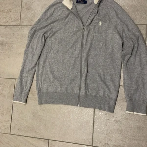 Grå kofta från Polo Ralph Lauren - Grå kofta från Polo Ralph Lauren med hel dragkedja och liten vit logga på bröstet. Tröjan har ribbade muddar och krage i kontrasterande vitt. Tillverkad i mjuk bomullsmix, perfekt för lager-på-lager under kyliga dagar.