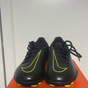 Nike Phantom svart/gul fotbollsskor - Snygga Nike Phantom fotbollsskor i svart med gula detaljer och blå text. Skorna har snörning, platt sula med dobbar och är tillverkade i syntetmaterial. Perfekta för dig som vill sticka ut på planen med stilrena och sportiga skor.