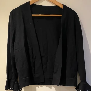 Svart kofta med volangärm och nitar. Aldrig använd. - Svart kofta med öppen front och långa ärmar. Ärmsluten har volangdetalj och är dekorerade med små nitar längs kanten. Koftan är stickad och har en klassisk, enkel design med en twist på ärmarna.