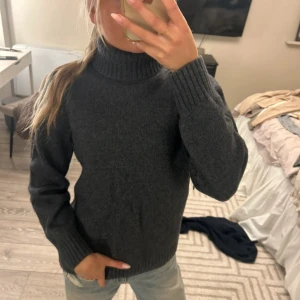 Mörkgrå polotröja i ull - Mysig mörkgrå polotröja med ribbade muddar och hög krage. Tröjan är stickad i ull och har en klassisk, enkel design som passar perfekt till jeans eller kjol. Perfekt för kyliga dagar när du vill vara både varm och snygg.