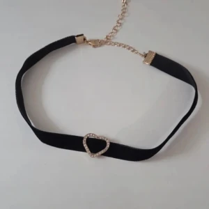 Svart choker med hjärthänge - Snygg svart choker med ett glittrigt hjärthänge i guld och strass. Halsbandet har en bred sammetsliknande rem och justerbart guldfärgat spänne. Perfekt accessoar för att lyfta din outfit med lite extra bling.