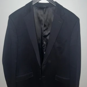 Mörklbå  klassisk kavaj - En stilren svart kavaj med klassisk passform och snygga slag. Kavajen har tre fickor framtill och är helfodrad med slät insida. Perfekt för dig som vill ha en tidlös och enkel look. Passar lika bra till skjorta som t-shirt.