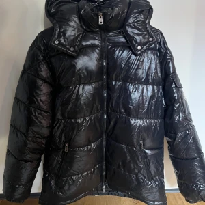 Svart Moncler Maya  - Snygg svart pufferjacka från Moncler i glansigt material. Jackan har huva, dragkedja framtill och två fickor med dragkedja. Klassisk Moncler-logga på ärmen och coolt innerfoder med serietidningsmotiv. Perfekt för kalla dagar.