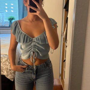Croppad topp från Zara - Säljer en turkos croppad topp från Zara med v-ringning, volangdetaljer och snörning framtill. Toppen har korta ärmar och är tillverkad i ett glansigt, ribbat syntetmaterial som ger en cool look. Helt ny, aldrig använd.