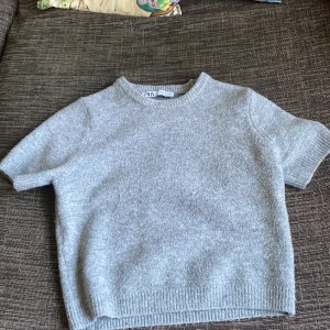 Grå stickad kortärmad tröja från Zara - Supermjuk och stilren grå stickad tröja från Zara med korta ärmar och rund hals. Tröjan har en enkel och clean look med ribbade kanter och passar perfekt till jeans eller kjol. Snygg och enkel att styla för en chill vibe.