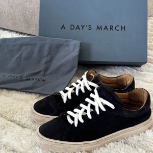 Svarta mockasneakers från A Day's March - Snygga svarta sneakers från A Day's March i mocka med vita snören och beige innersida. Klassisk låg modell med platt sula och rund tå. Perfekta för dig som gillar stilrena och tidlösa skor med en modern touch.