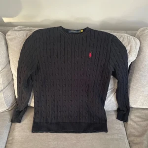 Svart kabelstickad tröja Polo Ralph Lauren - Svart kabelstickad tröja från Polo Ralph Lauren med röd broderad logga på bröstet. Klassisk rund halsringning och ribbade muddar vid ärmslut och nederkant. Perfekt för dig som gillar stilren och tidlös design.