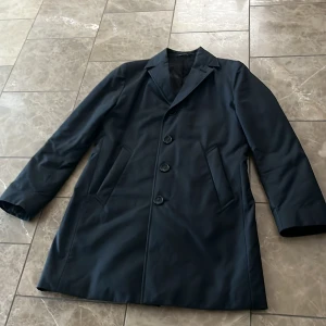 Svart rock från Tiger of Sweden, stl 50 - Stilren svart rock från Tiger of Sweden i klassisk modell med tre knappar framtill och snedställda fickor. Yttermaterial i bomull och polyamid, helfodrad med viskos. Perfekt för dig som vill ha en clean och tidlös look.