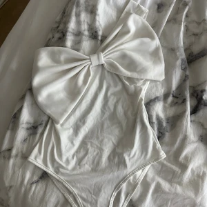 Vit body med stor rosett - Supersnygg vit body med en stor rosett framtill och asymmetrisk axel. Stilren och modern design som verkligen sticker ut. Fint skick förutom en liten defekt längst ner där man knäpper bodyn (kolla sista bilden) det är mina byxor som har färgat av sig🥰