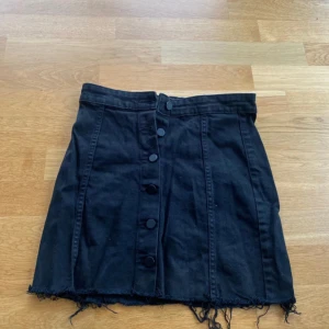 Svart jeanskjol med knappar - Svart kort jeanskjol med hög midja och fransig nederkant. Kjolen har knappar framtill och markerade sömmar. 