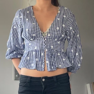 Blårutig blus med volang och broderi - Supersöt blus i blåvitt rutigt mönster med små vita broderade blommor. Djup v-ringning, knappar framtill och volang nertill. Lätt puffiga ärmar i trekvarts längd. Perfekt för en somrig look!