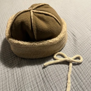 Beige och brun fleece mössa - Mysig mössa i beige och brun fleece med uppvikt kant och dekorativa sömmar. Tillverkad i 100% polyester. Perfekt för kalla dagar när du vill hålla dig varm och snygg.