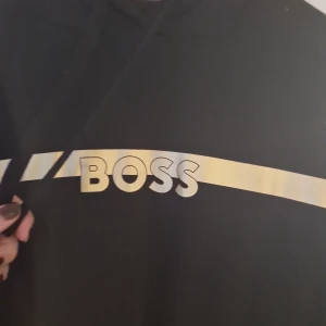 Svart hoodie från BOSS med gulddetalj - Svart hoodie från BOSS Homewear med en bred guldfärgad rand och BOSS-logga över bröstet. Tröjan har huva med snörning och långa ärmar. Perfekt för en chill och stilren look. Materialet är mjukt och skönt, troligen bomullsmix.