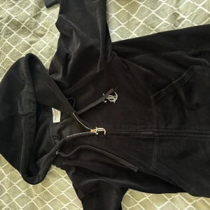 Svart Juicy Couture hoodie i velour - Svart hoodie från Juicy Couture i mjuk velour med glittrande Juicy-text i strass på ryggen. Tröjan har huva, dragkedja framtill, fickor och snygga gulddetaljer på snörena. Perfekt för dig som vill ha en ikonisk och trendig look.