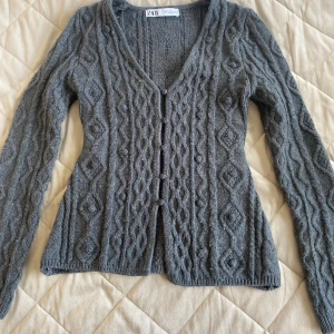 Grå stickad kofta från Zara - Mysig grå stickad kofta från Zara med snyggt kabelstickat mönster och v-ringning. Koftan har lång ärm och knäpps framtill med matchande stickade knappar. Perfekt för lager-på-lager och chill höst- och vinterstil.