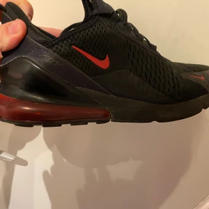 Nike Air Max 270 svarta/röda sneakers - Nike Air Max 270 sneakers i svart med röda detaljer och synlig röd air-bubbla i hälen. Ovandelen är i mesh med  ventilerande hål och klassisk Nike-logga på sidan. Skorna har snörning och en sportig, modern look som sticker ut. Skorna är i bra skicka men endast lite smutsiga på bilden. 