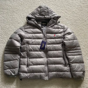 Grå dunjacka från Polo Ralph Lauren - Snygg grå dunjacka från Polo Ralph Lauren med huva och dragkedja framtill. Jackan har två sidofickor med dragkedja och den klassiska röda Polo-loggan broderad på bröstet. Perfekt för kyliga dagar och riktigt skön att bära. Jackan är aldrig använd och har inga anmärkningar. Tveka inte att höra av er vid några frågor eller funderingar 🤗