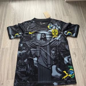 Svart Brasilien fotbollströja  - Unik svart Brasilien fotbollströja från Nike med coolt grafiskt tryck av Kristusstatyn och brasilianska symboler. Tröjan har gula detaljer, Brasil-logga och Nike Swoosh på bröstet. Tillverkad i Dri-FIT-material som andas och är perfekt för match eller träning.