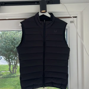 Mörkblå  dunväst från UBR, strl M - Mörkblå dunväst från UBR i storlek medium. Västen har quiltad design med horisontella sömmar, hög krage och dragkedja framtill. Materialet är polyester och dun, perfekt för kyliga dagar. Diskret UBR-logga på bröstet ger en clean look.
