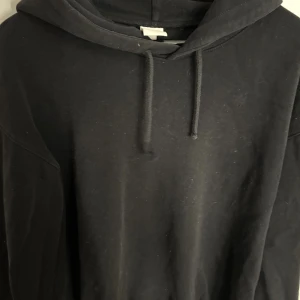 Svart hoodie  - En klassisk svart hoodie i mjuk bomull med huva och snörning. Perfekt för chill dagar eller när du vill ha en enkel och stilren look. Hoodien har normal passform och långa ärmar.