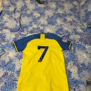 Al Nassr FC fotbollströja gul/blå - Gul och blå fotbollströja från Al Nassr FC med nummer 7 på ryggen och klubbmärke på bröstet. Tröjan har blå ärmar med gula detaljer och tryckt sponsorlogga framtill. Tillverkad i lätt och ventilerande material, perfekt för match eller träning. Ronaldo texten är borta på baksidan se bild på 