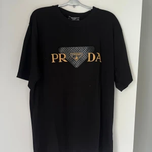 Svart Prada t-shirt med Paris-detalj - Svart t-shirt från Prada med guldfärgad brodyr och en quiltad, svart triangel med texten 'Paris' på bröstet. Klassisk rund hals och korta ärmar. Snygg och stilren design som sticker ut med exklusiva detaljer.
