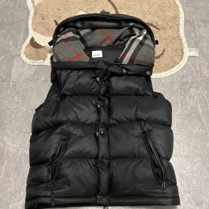 Svart dunväst från Burberry - Svart dunväst från Burberry med klassiskt rutigt foder i huvan. Västen har dragkedja och knappar framtill samt två fickor med dragkedja. Puffig och varm design, perfekt för kyliga dagar. Ikonisk Burberry-logga på insidan. Den är storlek m men kan passa s 170/92A 