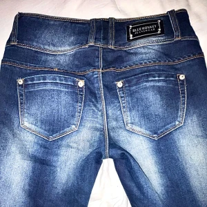 Blå jeans från Blue Monkey - Snygga blå skinny jeans från Blue Monkey med markerade slitningar och kontrastsömmar. tre knappar framtill ger en cool look. Klassisk femficksmodell och tydlig logga bak på linningen. 
