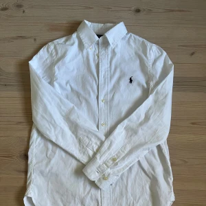 Vit skjorta från Polo Ralph Lauren - Klassisk vit skjorta från Polo Ralph Lauren i slim fit-modell. Skjortan har lång ärm, knappar framtill och ikonisk broderad logga på bröstet. Tillverkad i mjuk bomull och passar perfekt till en clean och stilren look. Står att skjortan är 16 men passar xs och är ungefär 164-170 cm. 