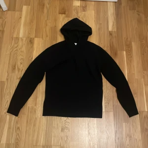Svart hoodie i merinoull från Vailent - Svart hoodie från Vailent i mjuk merinoull. Klassisk modell med huva och långa ärmar, perfekt för dig som gillar stilrena och bekväma plagg. Snygg att bära till jeans eller dressade byxor.